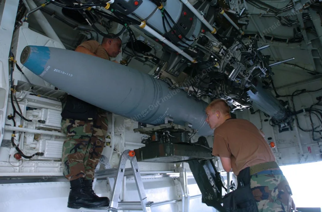 Two U.S. Air Force personnel are seen loading a large ordnance, likely a bomb, into the internal weapons bay of a B-2 Spirit stealth bomber. The aircraft’s intricate mechanical and hydraulic bomb-release system is visible above them, along with various wiring and structural components inside the bay. Both individuals are wearing camouflage pants and brown shirts, with one standing on a step ladder during the operation.