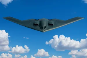 A high-resolution image of a Northrop B-2 Spirit stealth bomber flying against a vivid blue sky with scattered white clouds, showcasing its sleek, angular, flying-wing design.